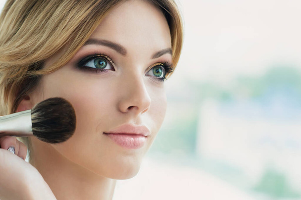 Model with bright color make up with long lashes and fresh young skin. Beautiful young woman with professional makeup and cosmetic powder brush in hand front of the window.  Close up, copyspace. - Foto, imagen