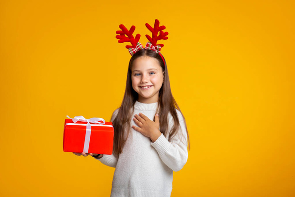 Καλές γιορτές. Χαμογελαστό κορίτσι με κέρατα σε πουλόβερ κρατά δώρο στο χέρι - Φωτογραφία, εικόνα