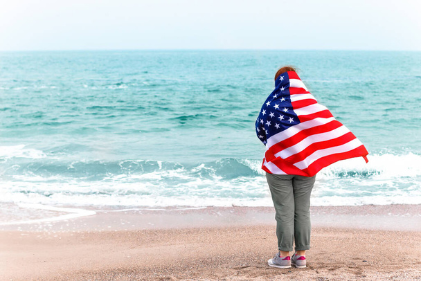 Pohled zezadu na mladou dívku s americkou vlajkou hledí na moře - Fotografie, Obrázek