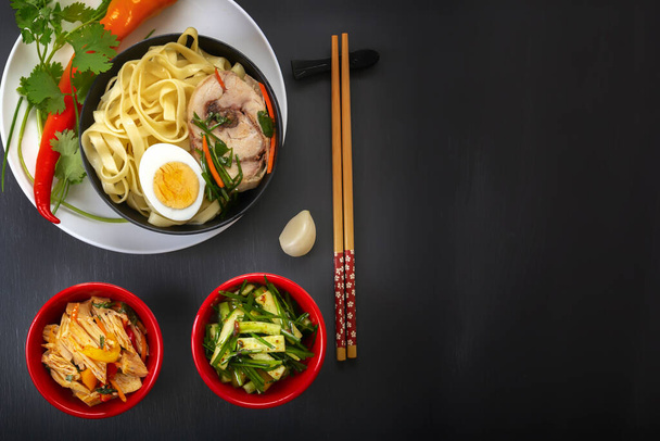 Noodles with pieces of meat and egg in a black plate and dim sums with different traditional snacks of mushrooms, soy and vegetables with chopsticks on a dark table. View from above. Asian food concept. Copy space - 写真・画像