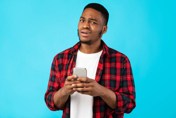 Hombre negro inseguro sosteniendo el teléfono celular posando sobre fondo azul - Foto, Imagen