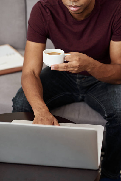 Przycięty obraz młodego czarnego człowieka pracującego na laptopie i pijącego kawę - Zdjęcie, obraz