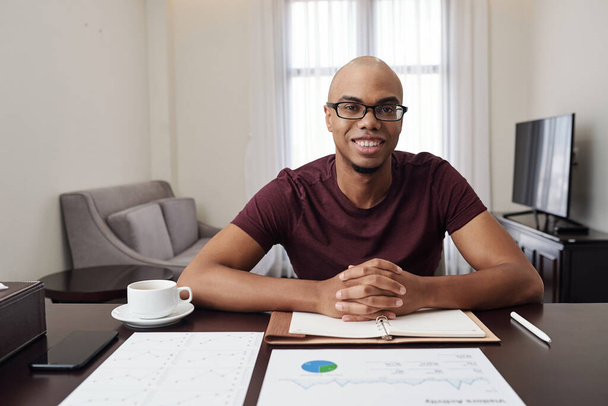 Портрет красивого улыбающегося молодого чернокожего бизнесмена в очках, сидящего за столом дома и работающего с финансовыми документами - Фото, изображение