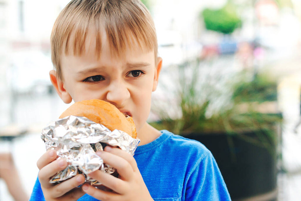 空腹の少年は屋外のカフェでバーガーを食べる。かわいい子供はファーストフードを食べる。子供の頃、不健康な食品の概念。ジャンキー・フード。生活と人. - 写真・画像
