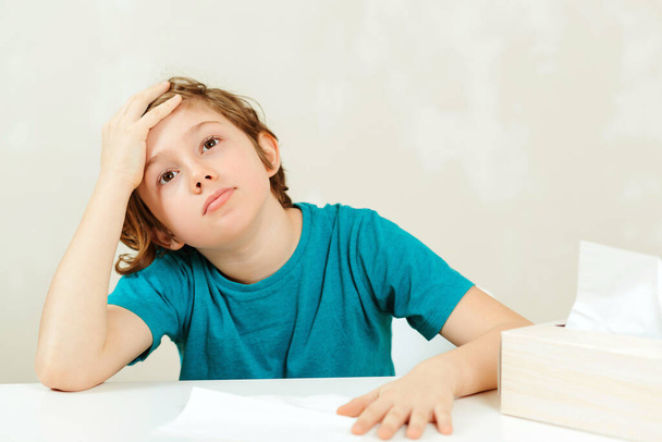 Schoolboy has headache during lessons. Sick boy sitting at the desk. Boy has a virus, runny nose and headache. Healthcare and childhood. - 写真・画像