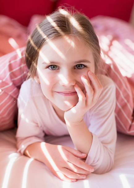 Cute little girl in bed woke up in the morning. Child lies in bed and holds hands under cheek. The rays of the sun through the blinds on the kid's face. Pastime at home, comfort. Carefree childhood - Photo, Image
