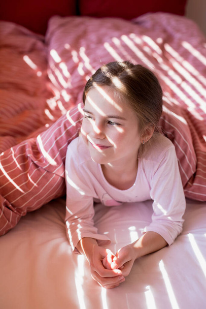 Cute little girl in bed woke up in the morning and looks towards the window. The sun's rays fall on the child's face through the blinds. Pastime at home. Home comfort. Carefree childhood - 写真・画像