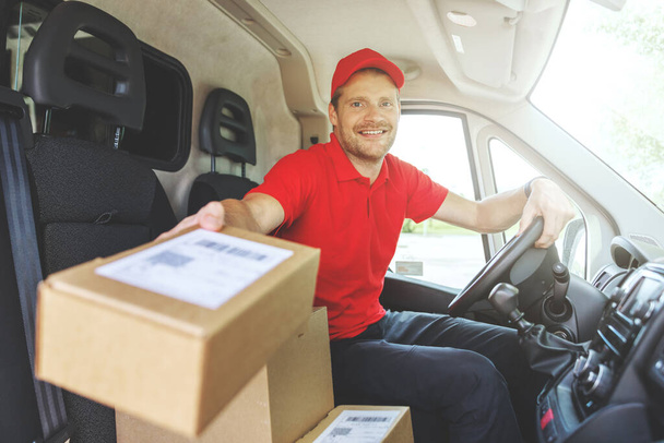 fiatal mosolygós kézbesítő férfi ül a furgonban és dobozokat szállít. a csomag megadása - Fotó, kép