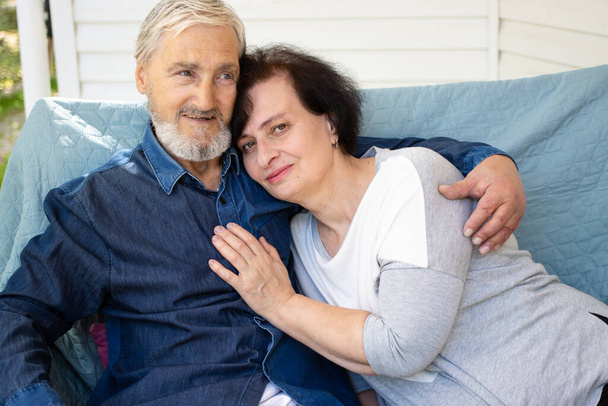 Happy mature husband and elderly wife relaxing sitting on sofa at home terrace, hugging and cuddling, smiling senior loving couple spending tender romantic weekend together. Showing care affection - Photo, Image