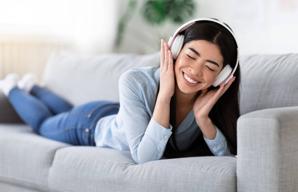 Time To Relax. Cheerful Asian Girl In Headphones Listening Music On Couch - Foto, afbeelding