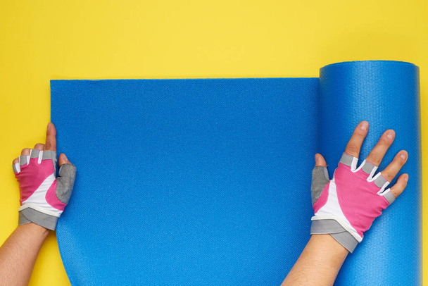 two female hands in sports gloves unfold a blue yoga mat, top view, yellow background  - Photo, Image
