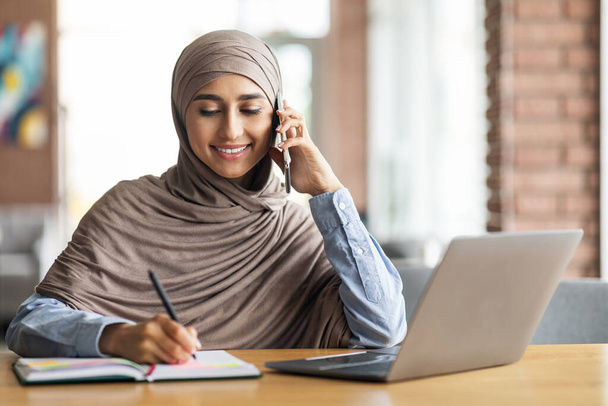 Young woman in hijab talking on phone, taking notes - Foto, Imagem