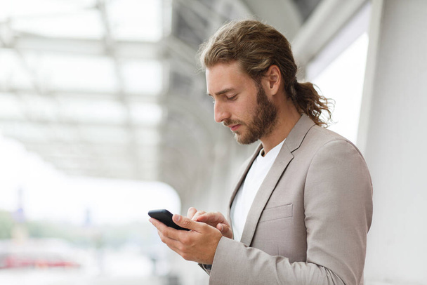 Portrait of handsome curly businessman in casual wear holding smartphone and smiling.Successful manager using mobile phone apps,texting message,browsing internet,looking at phone near business center - Foto, immagini