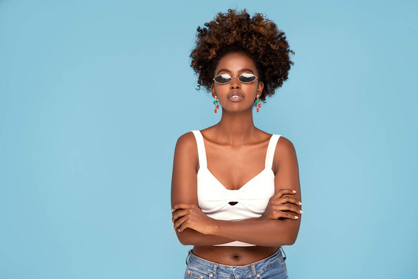 Fashionable young african girl with afro hairstyle posing in studio, wearing sunglasses and earrings. Blue pastel studio background. - Fotó, kép