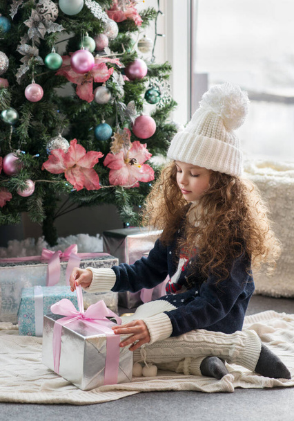 Cute little girl in winter clothes posing in home front of Christmas tree with gift box in hand. X-mass and New year waiting and celebration concept. Holiday home decoration. - Fotoğraf, Görsel