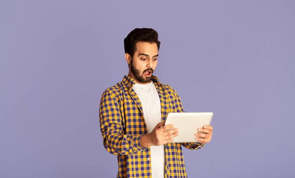 Shocked Indian man staring at tablet screen with open mouth on violet background - Фото, изображение