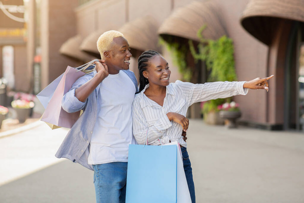 Glückliches junges Paar mit Einkaufstüten spaziert, Frau zeigt mit dem Finger auf Schaufenster - Foto, Bild