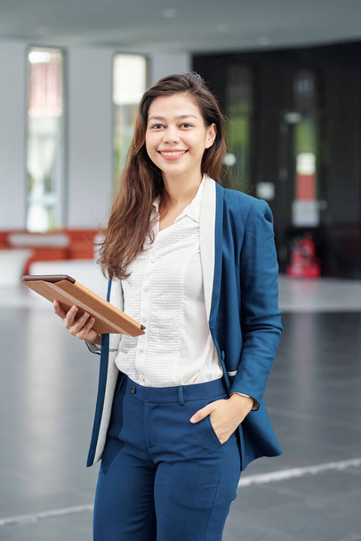 Портрет прекрасной молодой бизнес-леди, стоящей в офисном зале с цифровой табличкой в руках - Фото, изображение