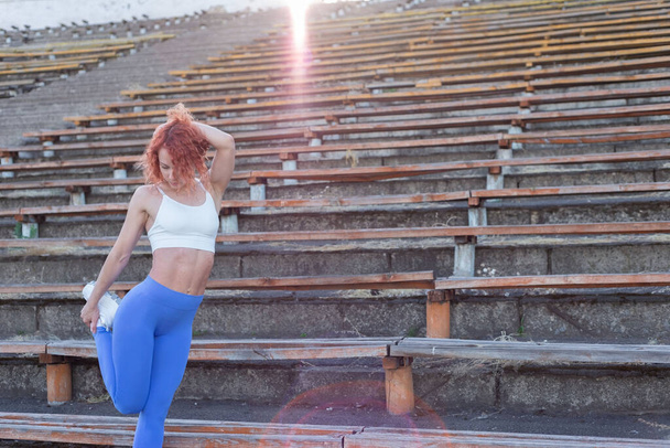 portrait of a red-haired female athlete, thirty-six years old, in a turquoise sports uniform, doing a warm-up at a sports stadium. active lifestyle, sports and health. Outdors Motion of fit woman stretching upper body before training at stadium in su - Photo, Image