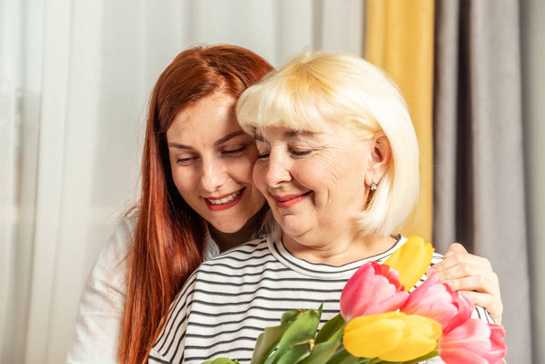 Доросла блондинка тримає букет тюльпанів, а її дочка обіймає будинок. Щасливий день матері. - Фото, зображення
