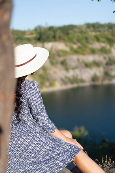 uma menina de chapéu e vestido, sentada ao lado de uma árvore no topo de uma montanha, olha para a distância no lago. menina na natureza durante o pôr do sol, Relaxe o tempo em viagens conceito de férias, foco seletivo e suave - Foto, Imagem