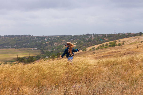 Schattig klein meisje in spijkerjasje, zwarte zonnebril, blauwe ruitjurk in geel grasveld. Gelukkige stijlvolle lange blonde haar kind op het platteland landschap. Schattig kind wandelen outdoor landelijke road trip. - Foto, afbeelding