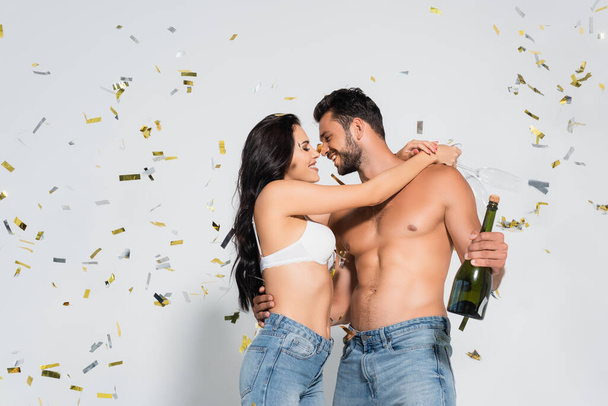 brunette woman in bra hugging muscular man with bottle of champagne on grey with confetti  - Photo, Image