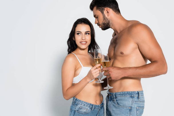 muscular man looking at sexy woman in bra holding glass of champagne on grey - Photo, Image