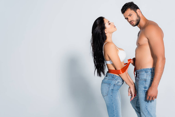 muscular and bearded man tying sexy woman in bra and jeans on white - 写真・画像