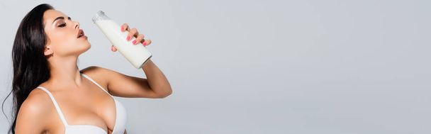 panoramic crop of sexy woman in bra holding bottle with milk isolated on grey - Foto, Imagem