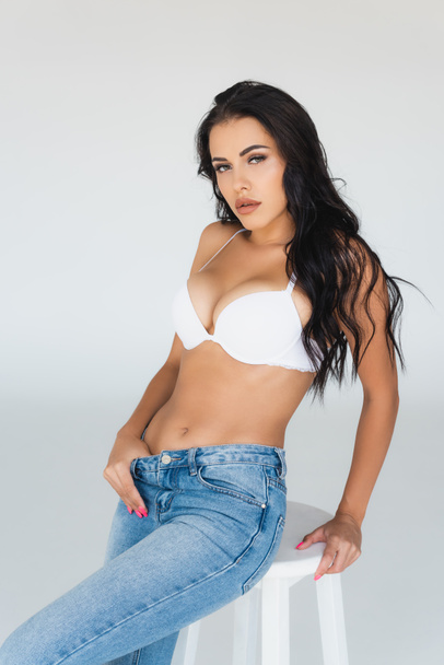 sexy young woman in bra and blue jeans sitting on stool and looking at camera on grey - Foto, Imagem