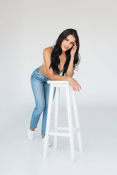 sexy woman in bra and jeans leaning on stool and looking at camera on grey - Fotografie, Obrázek
