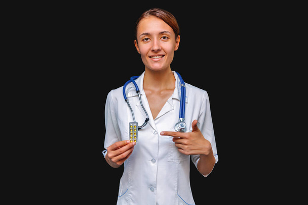 Usmívající se doktorka ukazuje prstem na prášky. Izolováno na šedém pozadí - Fotografie, Obrázek