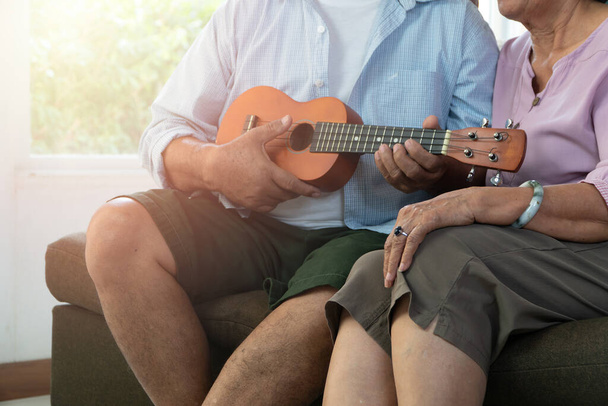 幸せなアジアの高齢者のカップルがウクレレを演奏し、自宅で一緒に歌う。幸せな引退後の高齢者のための生活の概念 - 写真・画像