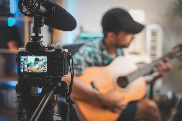 Behind the scene shot of a young man influencer making a video playing guitar. Very handsome man with beard shooting a video for social media talking directly to the camera. Influencer's work space. - Zdjęcie, obraz
