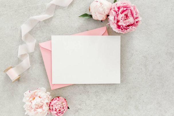Mockup invitation, blank paper greeting card, pink envelope and peonies on gray stone table. Flower background. Flat lay, top view. - Foto, Bild