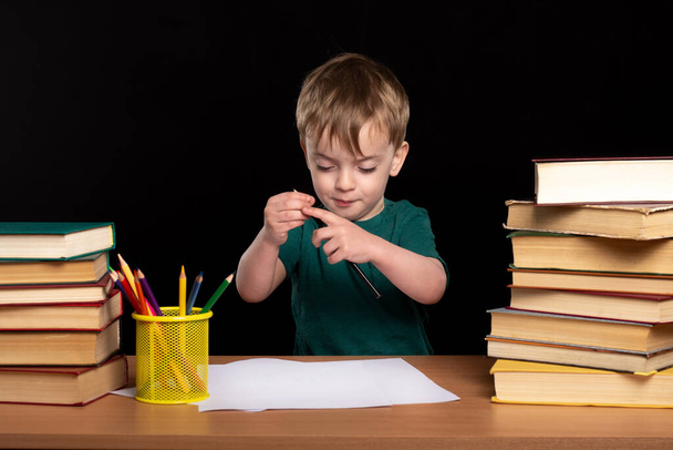 3歳の少年が本を持ってテーブルに座り、鉛筆で絵を描く。隔離中の遠隔学習。子供は宿題をする。ヨーロッパの外観. - 写真・画像