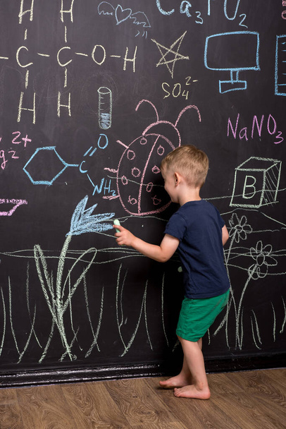 bambino di tre anni disegna con gesso multicolore su una grande lavagna. parete nera con disegni e formule per bambini. fotografia verticale. - Foto, immagini