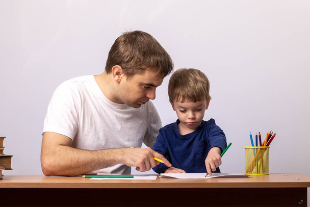 Egy kisfiú apával leül egy asztalhoz könyvekkel, és nézi, hogyan kell írni. Szürke otthoni kényelmi háttér. családi kikapcsolódás. tanár megtanítja a gyermeket rajzolni.  - Fotó, kép