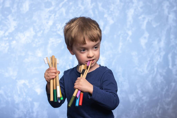 小さな男の子がバスルームでたくさんの竹の歯ブラシを保持し、検査します。生分解性口腔衛生用品。環境に優しい. - 写真・画像