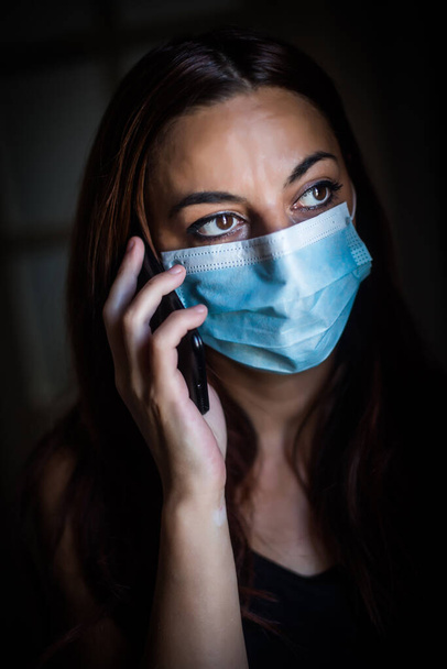 Portrait of a young woman talking on the mobile phone while wearing a mask, indoor, by a window. - Photo, Image
