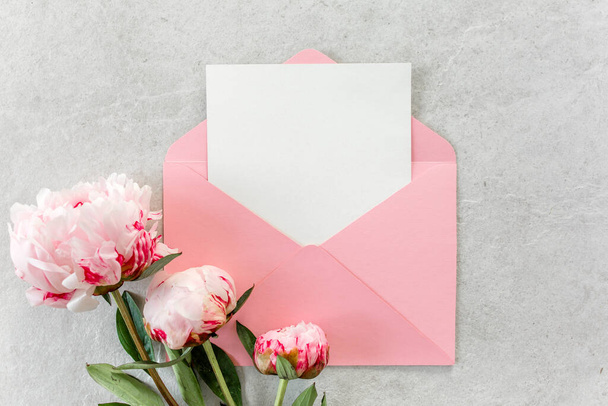 Mockup invitation, blank paper greeting card, pink envelope and peonies on gray stone table. Flower background. Flat lay, top view. - Photo, image