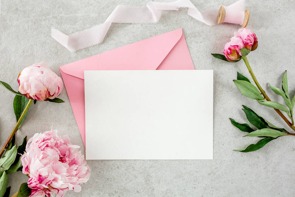Mockup invitation, blank paper greeting card, pink envelope and peonies on gray stone table. Flower background. Flat lay, top view. - Foto, imagen