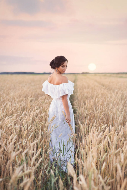 Portrait of a young beautiful brunette girl in a white summer dress standing in a field with wheat ears on the background of the sunset sky - Photo, image