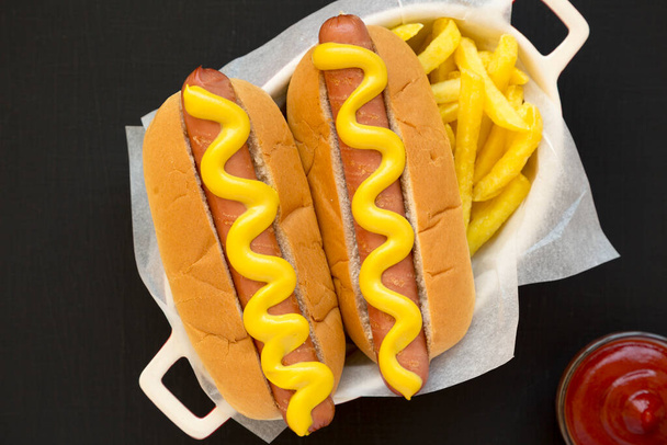 Homemade Mustard Hot Dog, French Fries on the black surface, top view (англійською). Плоть лежала, вгорі, зверху..  - Фото, зображення