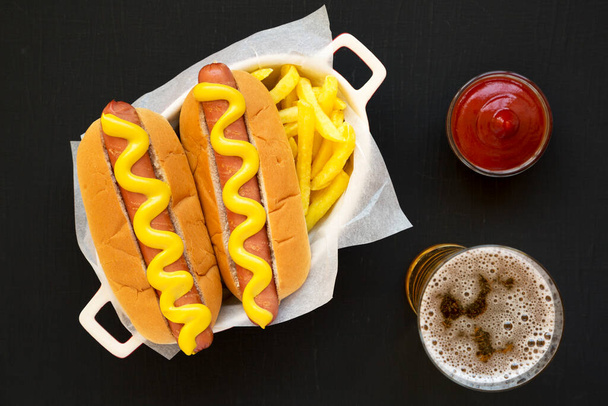 Homemade Mustard Hot Dog, French Fries and Glass of Beer on a black background, overhead view. Flat lay, top view, from above. - Photo, Image