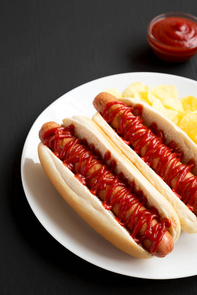Maukas amerikkalainen hot dog perunalastujen kanssa valkoisella lautasella mustalla pinnalla, sivunäkymällä. Tekstitila. - Valokuva, kuva