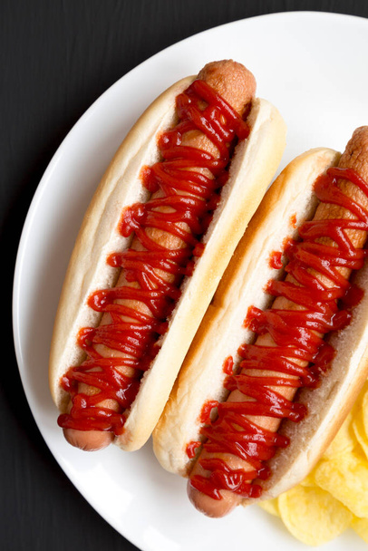 Вкусный американский Hot Dog с картофельными чипсами на белой тарелке на черном фоне, вид сверху. Плоский лежал, сверху, сверху. - Фото, изображение