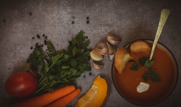 Pumpkin soup in a bowl with crackers, with fresh pumpkins, garlic and parsley, carrots, tomatoes, on a dark background. view from the top, tint. - Photo, Image