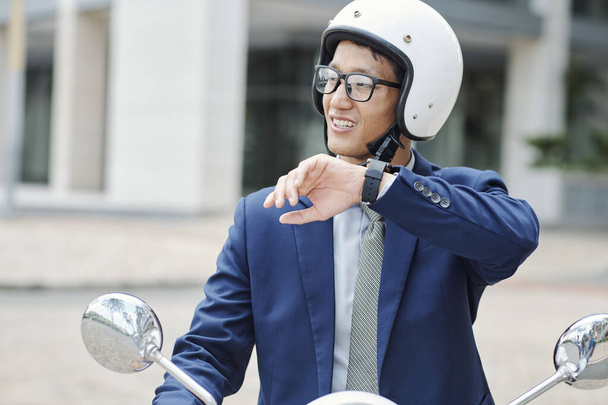 Porträt eines jungen lächelnden Geschäftsmannes mit Helm, der auf einem Roller fährt und Sprachnachrichten per Anwendung auf der Smartwatch aufzeichnet - Foto, Bild
